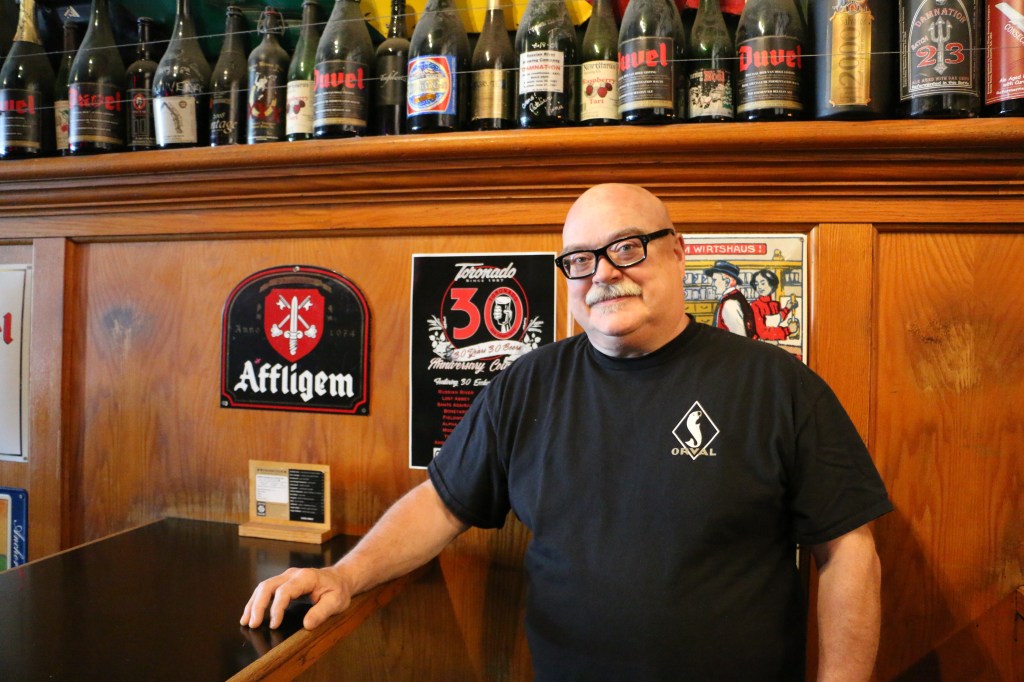 Photo of Dave Keene wearing a black Orval tee shirt in 2017, inside the Toronado next to a 30th Anniversary Poster and an Affligem Belgian Beer plaque.