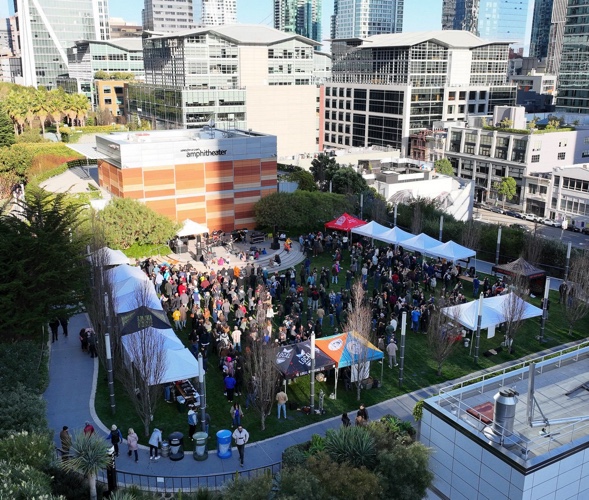Drone view of rooftop park with skyscrapers surrounding. Festival tents are being visited by hundreds of people.